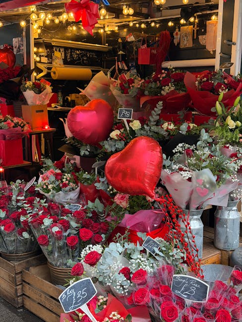 A vibrant floral display inside a florist shop, featuring an abundance of red and white flowers including roses, carnations, and sprigs of greenery. The arrangement is accented with three large red he